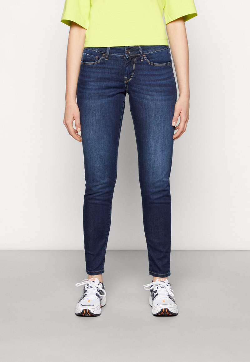 Jeans ajustados de mezclilla elástica en azul oscuro, con un diseño de cinco bolsillos, cremallera y costuras en contraste. Combinados con zapatillas blancas.