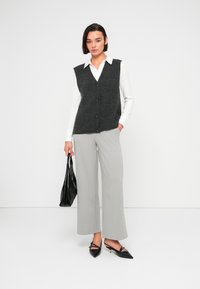 Γυναίκα που φοράει λευκό πουκάμισο με μακριά μανίκια, μαύρο πλεκτό γιλέκο με κουμπιά, ανοιχτό γκρι παντελόνι φαρδιάς γραμμής, μαύρες μυτερές ψηλοτάκουνες γόβες και κρατάει μαύρη τσάντα χειρός.