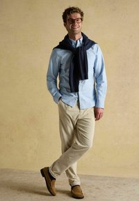 Usmievajúci sa muž v okuliaroch stojí uvoľnene, v svetlomodrej košeli, béžových kordových nohaviciach, hnedých mokasínach a tmavomodrom svetre prehodenom cez ramená.