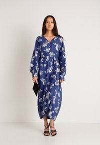 Μπλε φλοράλ μακρύ φόρεμα με μακριά μανίκια, λαιμόκοψη V, σφιχτή μέση και φουσκωτό φούστα. Συνδυασμένο με μαύρες λουρίδες σανδάλια και τσάντα clutch.