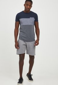 Navy and gray striped t-shirt with pocket, paired with gray shorts. Model wears black sneakers. Smooth fabric with relaxed fit.