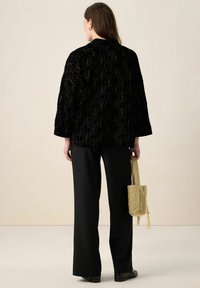 Zwarte fluwelen jas met geometrisch patroon, losse pasvorm, driekwart mouwen; gecombineerd met zwarte broek met wijde pijpen en een kleine gouden tas.