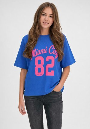 Jonge vrouw met lang bruin haar die een blauw Miami City 82 T-shirt en zwarte spijkerbroek draagt, staand met één hand in de zak.