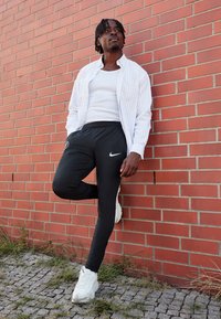 Melnas sporta bikses ar ribām un baltu Nike logo, saskaņotas ar baltu bezrūpīgu krekliņu un svītrainu baltu krekliņu ar pogām. Izskatu papildina baltas kedas.