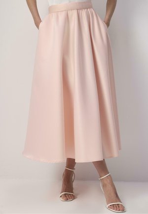 Frau trägt einen hellrosa A-Linien-Rock in mittlerer Wadenlänge mit Taschen und weiße, offene Sandalen mit Knöchelriemen, steht vor einem schlichten Hintergrund.