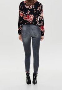 Mujer con una blusa negra de mangas largas con flores, jeans ajustados grises y botas de tacón negras, de pie de espaldas a la cámara.