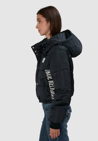 Czarna kurtka puchowa z kapturem, pikowana tekstura, zamek z przodu i szczegół z białym logo z boku. Nosi się ją z niebieskimi dżinsami.