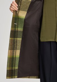 Hand hält einen grünen und braunen karierte Mantel offen, der das dunkle, glatte Innenfutter zeigt, getragen über einem olivgrünen Oberteil und marineblauen Hosen.