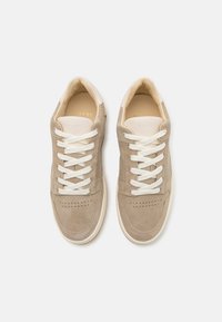 Beige suede lenkkarit, joissa on valkoiset nauhat, kärjessä rei'ityksiä ja valkoinen kumipohja. Minimalistinen muotoilu, jossa on hienovarainen brändäys.