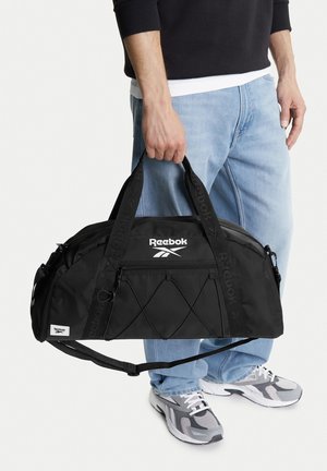 Mann hält schwarzen Reebok-Reisetasche an den Griffen, trägt hellblaue Jeans, schwarzes Langarmshirt und graue Turnschuhe.