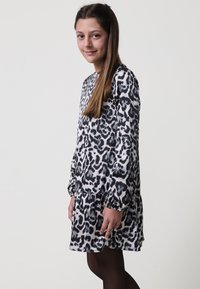 Haljina s uzorkom leoparda u crnoj i bijeloj boji, dugih rukava, nabranog rubnog dijela, lepršavog materijala, nosi se s crnim tajicama.