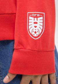 Μανίκι κόκκινης φανέλας με το λευκό λογότυπο ÖFB που απεικονίζει έναν στυλιζαρισμένο αετό και μια ποδοσφαιρική μπάλα, φορεμένο πάνω από μπλε τζιν παντελόνια.