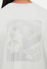 Parte trasera de una camiseta blanca con un sutil diseño geométrico gris que presenta un balón de fútbol con patrón y líneas verticales.