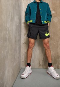 Person som bär en teal jacka med gula knappar, svarta Nike-shorts med neonswoosh, svarta strumpor och vita Nike-sneakers står mot betongväggar.