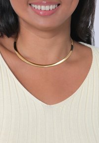 Goldfarbene Metall-Choker-Halskette mit einem geschwungenen, flachen Design; ausgestattet mit einem Kettenverschluss, getragen über einem hellen, strukturierten Oberteil.