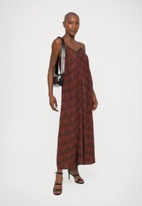 Vestido maxi padrão vermelho e castanho com um ajuste solto, alças finas e um design fluido. Modelo carrega uma mala preta e usa sandálias de tiras.