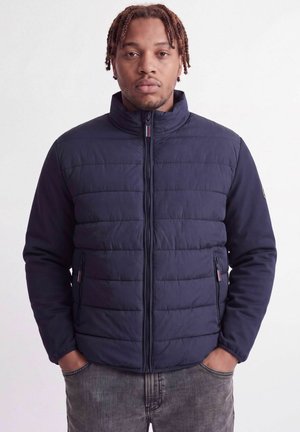 Homme aux cheveux tressés portant une veste matelassée bleu marine zippée et un jean gris, mains dans les poches, sur un fond uni.