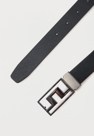 Μαύρη δερμάτινη ζώνη με υφή, με ασημένια ορθογώνια αγκράφα που διαθέτει μοντέρνο γεωμετρικό σχέδιο κοπής, ακουμπισμένη επίπεδα σε λευκή επιφάνεια.