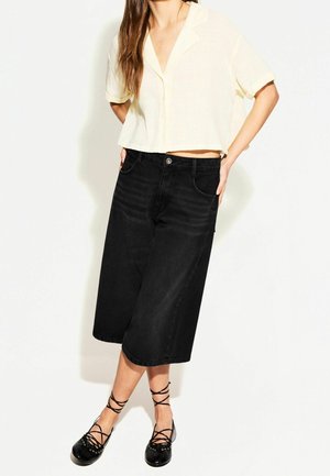 Femme portant un haut court crème à manches courtes et boutons, un pantalon large noir coupé à la cheville, et des ballerines noires à lacets sur fond blanc.