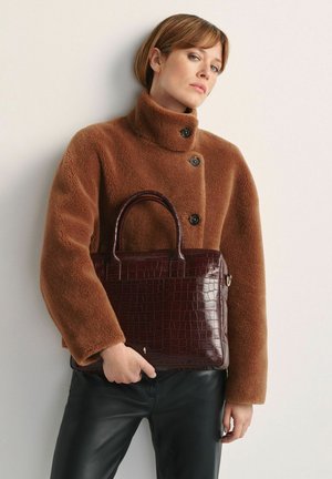 Kobieta w brązowej, zapinanej na guziki polarowej kurtce i czarnych spodniach trzyma ciemnobrązową torebkę o fakturze krokodyla na tle białej ściany.