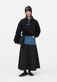 Sudadera negra con cremallera y logo, chaleco de mezclilla azul, falda larga negra, bolso negro, gorro de canalé negro y zapatos negros.