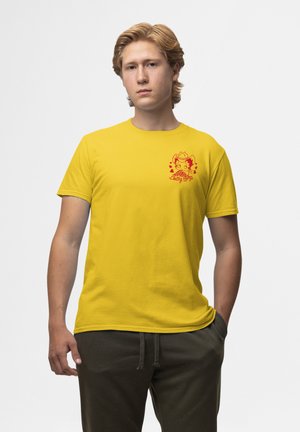 Gul bomulds t-shirt med korte ærmer. Har et rødt grafisk design på venstre bryst, der viser en karakter og tekst. Glat tekstur.