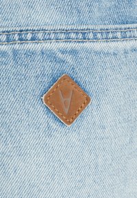 Tecido de denim em azul claro com uma superfície texturizada. Apresenta um retângulo em forma de diamante em couro castanho com um logótipo em relevo e costura contrastante.