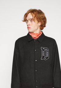 Black jacket featuring a button closure and side pockets, accented with a white embroidered logo on the chest. Bandana in orange around the neck.