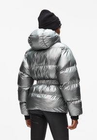 Giacca puffer argento con cappuccio, caratterizzata da una finitura lucida, vita elastica e polsini a coste neri. Cuciture dettagliate sulle spalle.