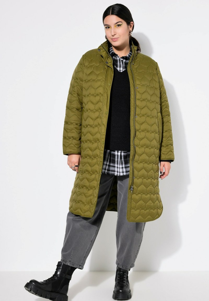 Cappotto trapuntato verde oliva con colletto alto, caratterizzato da un design gonfio e un motivo a forma di cuore, indossato sopra un maglione nero e una camicia a scacchi.