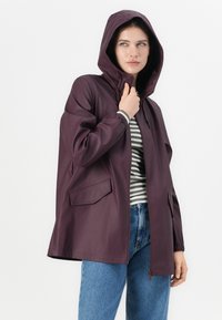 Burgunderröd huvtröja i vattentåligt material med elastiska manschetter, en framdragkedja och två sidofickor. Bärs över en randig topp och jeans.