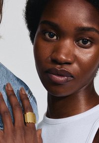 Bague en or texturée avec des stries verticales portée sur la main. La peau est d'un brun profond ; la chemise est blanche. L'arrière-plan est neutre et discret.
