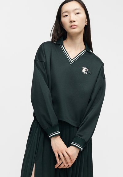 Jeune femme aux longs cheveux noirs porte un pull vert foncé à col avec des rayures blanches et une jupe plissée, se tenant devant un fond uni.