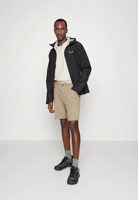 Sort vandtæt jakke med hætte, beige shorts, grå sokker og sorte sneakers. Jakken har en lynlåslukning foran og et logo på brystet.