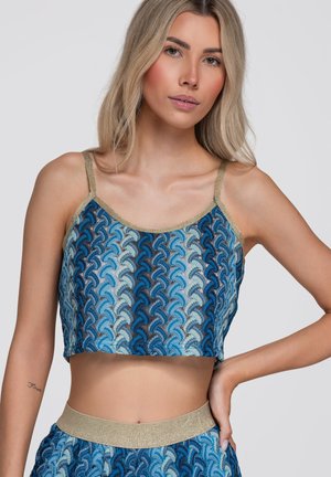 Ung kvinde iført en blå mønstret crop top med guldstropper og taljebånd, poserer med den ene hånd på hoften mod en ensfarvet baggrund.