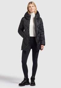 Manteau matelassé noir avec capuche, manches longues et fermeture éclair à l'avant. Porté sur un col roulé blanc, associé à des leggings noirs et des baskets.