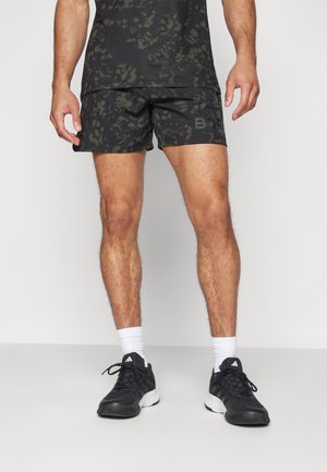Mężczyzna w czarnych i zielonych sportowych szortach z kamuflażowym wzorem oraz dopasowanej bluzce, białych skarpetkach i czarnych butach marki Adidas stoi na szarej podłodze.