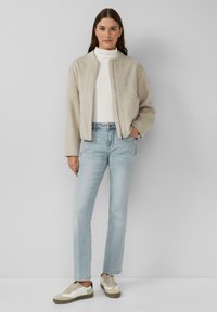 Helle beige Wolle Bomberjacke mit Reißverschluss, getragen über einem weißen Rollkragenpullover, kombiniert mit hellblauen geraden Jeans und beigen Sneakers.
