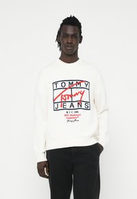 Cremfarbener Strickpullover mit einem auffälligen roten und blauen Logo-Design, auf dem "TOMMY JEANS" steht, mit gerippten Bündchen und einem klassischen Rundhalsausschnitt.