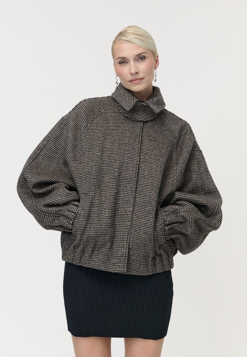 Жена със светла коса, носеща висококатова каре голяма яке и черна ребриста пола, стои пред бял фон.