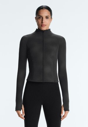 Žena stojaca čelom vpredu, oblečená v priliehavom čiernom dlhom rukáve so zipsom a čiernych nohaviciach na jednoduchom svetlom pozadí.