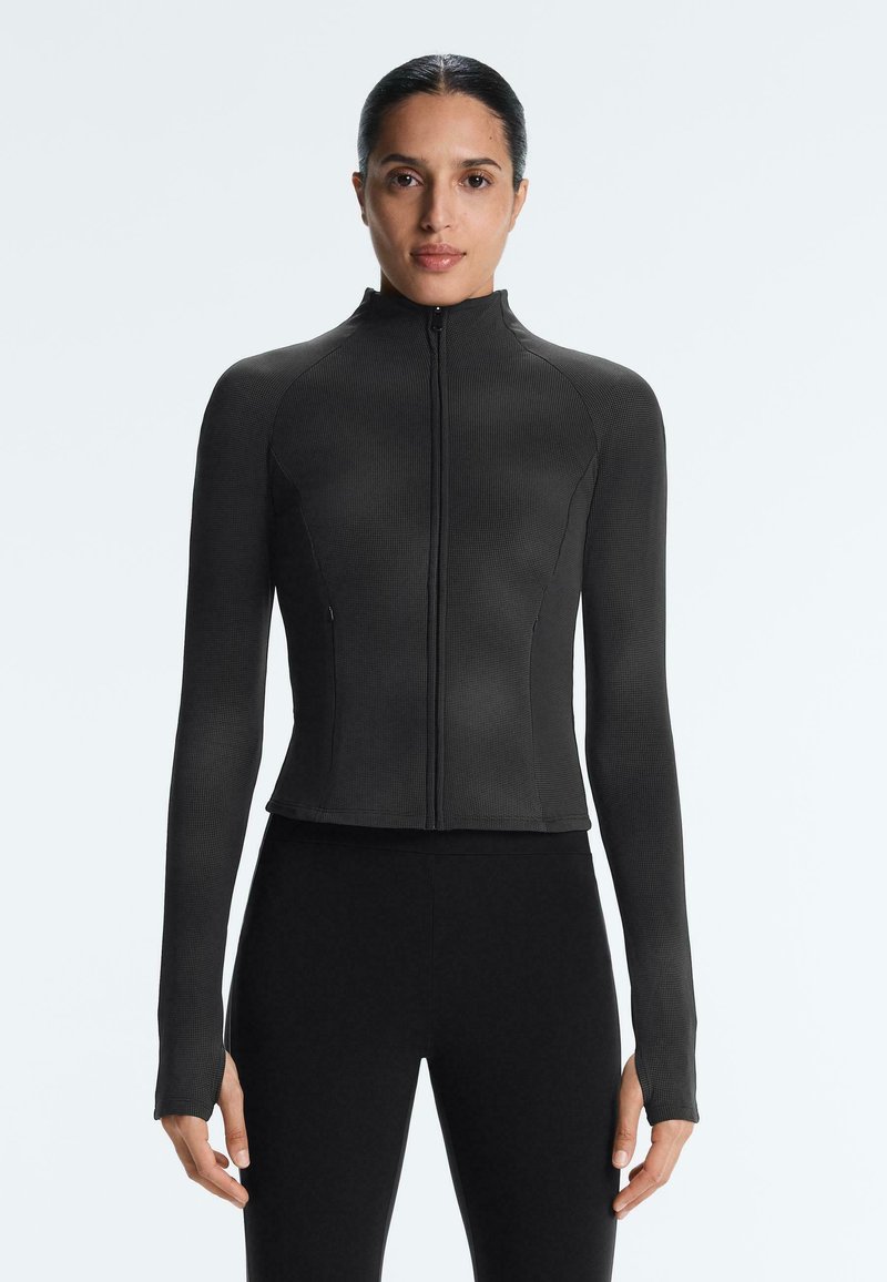 Femme debout, face à l'avant, portant un haut noir moulant à manches longues avec fermeture éclair et un pantalon noir, sur un fond clair uni.