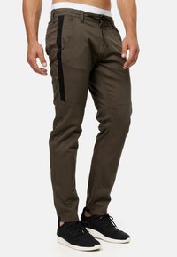 Olijfgroene broek met zwarte zijpanelen, taps toelopende pasvorm, elastische tailleband en trekkoorden. Draagt zwarte sneakers.