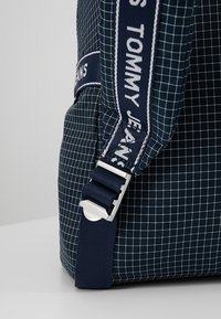 Mörkblå ryggsäck med rutmönster, utrustad med en vadderad axelrem, silverfärgad metallbeslag och en märkesvävd etikett. Hållbar tygtextur.