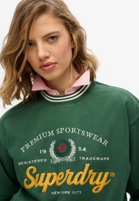 Sudadera verde con cuello a rayas blancas y rosas, con bordado blanco y amarillo: "Ropa deportiva premium" y "Superdry Nueva York".