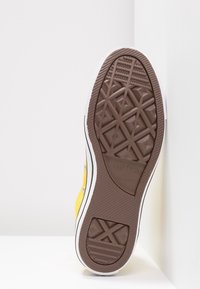 Gul lærredssneaker med brun gummisål, tekstureret mønster for greb, hvide accenter og metaløjer til snørebånd. Mærkelogo synligt.