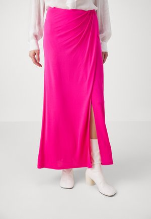 Fuchsia maxi skjørt laget av glatt stoff med en sideslitt, kombinert med hvite hælte ankelstøvler, som skaper en kontrast i farger og teksturer.