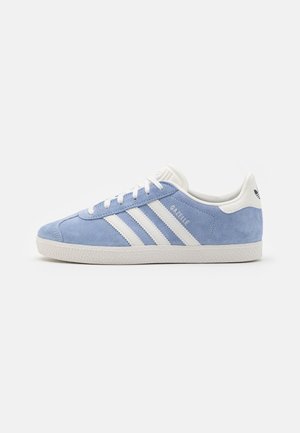 Baskets Adidas Gazelle bleu clair avec des bandes blanches, une semelle blanche et une languette blanche au talon sur un fond blanc uni.
