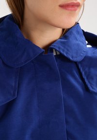 Blå bomullskappa med en stor krage, med en slät textur och en strukturerad design; inkluderar vertikala tygstrimmor inuti kragen.