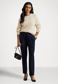 Camisola de malha creme com padrão de trança, calças tailored azul-marinho, saltos altos pretos e uma pequena mala preta com detalhes dourados.